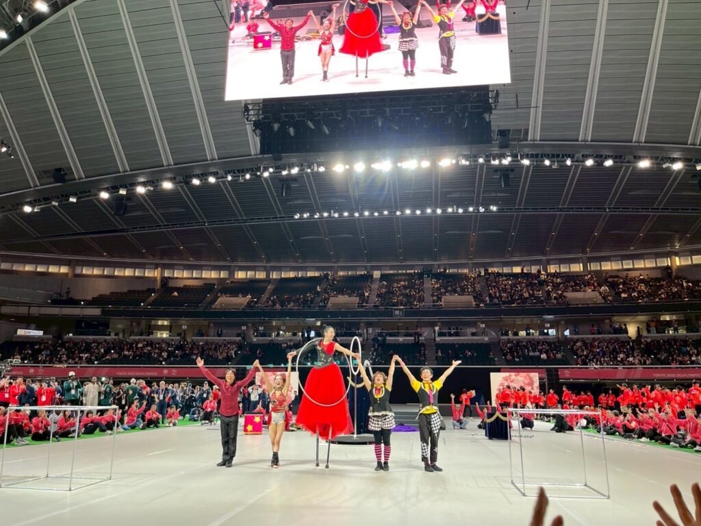 東京2025デフリンピック閉会式・式典前プログラム出演のヘブンアーティスト。左からJuggler Laby(ジャグリング)、めりこ(ポールアクロバットダンス)、AYUMI(フープ、スティルト)、Cartoon Clown CHEEKY! (一輪車サーカス)、しおじゃり(一輪車サーカス)。終演後の挨拶をするヘブンアーティストの周りを、赤いジャージのスタッフ、各国の選手団が囲んでいる。東京体育館にて。